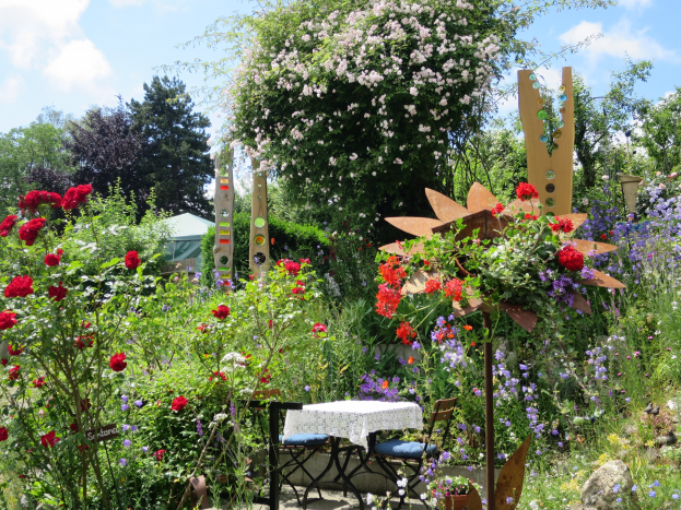 Ein Garten auf dem Chelsea Flower Show mit bunten Blumen und Pflanzen, einem Tisch mit einer Decke und Stühlen, umgeben von Bäumen unter einem bewölkten Himmel.