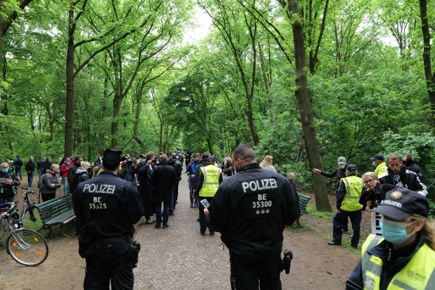 Polizisten vor einer Menschenmenge bei einer Outdoor-Veranstaltung, mit Fahrrädern und einer Bank im Vordergrund, Bäumen und Himmel im Hintergrund.