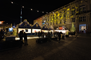 Ein lebendiger Weihnachtsmarkt in Berlin, Deutschland, mit Menschen um beleuchtete und dekorierte Stände, vor Gebäuden mit Fenstern unter einem dunklen Himmel.