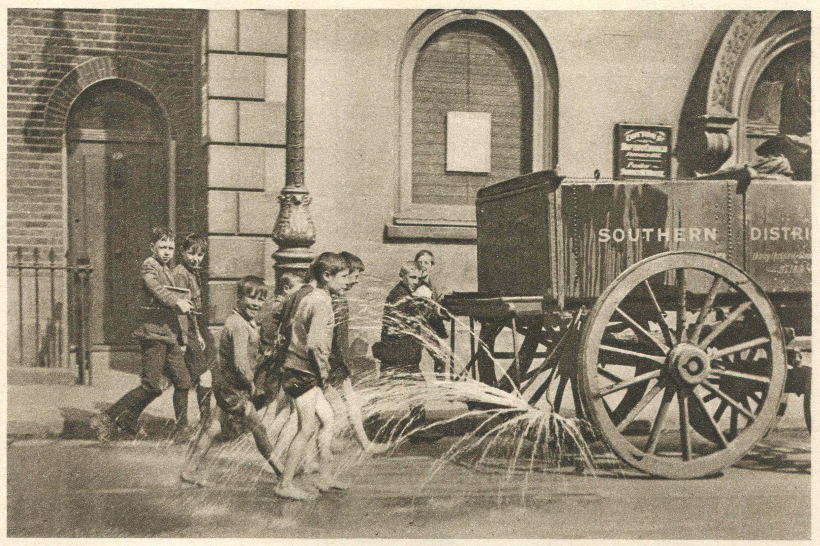 Schwarzes und wei├čes Bild von Kindern, die mit einem Feuerhydranten vor einem Geb├Ąude mit Fenstern, einer Tür, einem Mast und einem Zaun spielen, mit einem Wagen im Hintergrund.