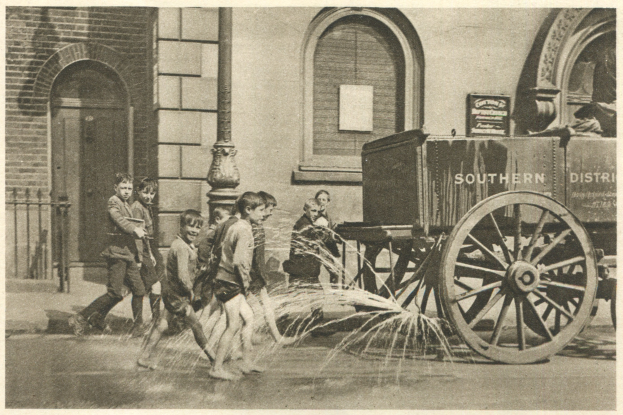 Schwarzes und wei├čes Bild von Kindern, die mit einem Feuerhydranten vor einem Geb├Ąude mit Fenstern, einer Tür, einem Mast und einem Zaun spielen, mit einem Wagen im Hintergrund.