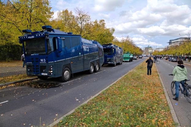 Eine Gruppe von Polizeifahrzeugen steht am Straßenrand mit einem Fahrradfahrer auf der rechten Seite, viele Menschen halten Fahrräder, Gras und trockene Blätter auf dem Boden und Bäume, Gebäude und ein bewölkter Himmel im Hintergrund.