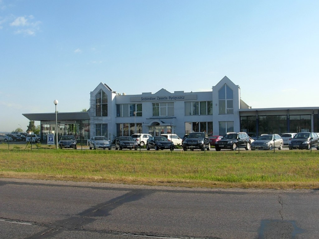 Ein Autohaus mit parkenden Autos vorne, umgeben von Gebäuden, Straßeninfrastruktur und Bäumen unter einem bewölkten Himmel.