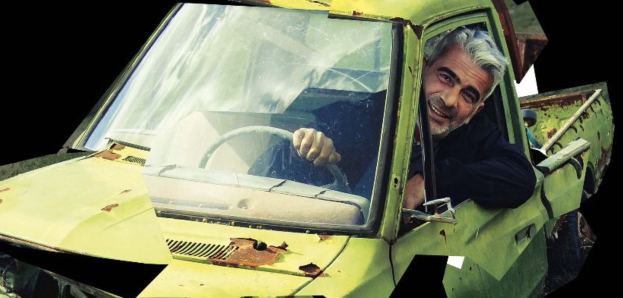Ein Mann lächelt, während er in einem gelben, rostigen und verbeulten Auto auf dem Fahrersitz sitzt und das Lenkrad hält.
