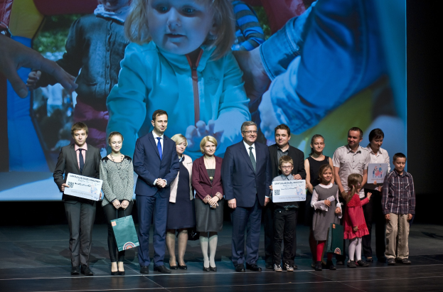 Gruppe von Menschen auf einer Buhne mit Taschen und Urkunden, mit einem Bildschirm im Hintergrund, der ein Kind und andere zeigt.
