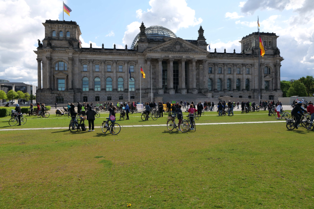 Eine Gruppe von Menschen, die vor dem Reichstaggebäude in Berlin, Deutschland, Fahrräder fahren, mit sichtbaren Fenstern, Säulen und Fahnenmasten des Gebäudes, umgeben von Bäumen und Gras unter einem bewölkten Himmel.