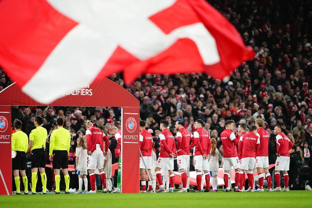 Eine Gruppe von Menschen auf einem Fußballfeld mit einer roten und weißen Flagge im Vordergrund, einem Bogen mit "Bayern München vs. Bayern München Wett-Tipps & Vorschau" im Hintergrund und einer großen Menge im Stadion.