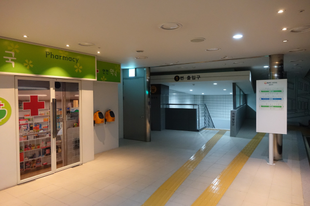 Apothekeneingang mit einem "Apotheke"-Schild links, einer Tafel mit Text rechts, beleuchteten Lichtern oben und Hintergrundobjekten.