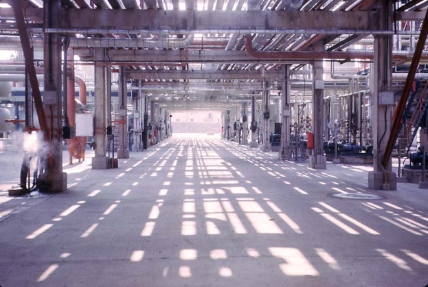 Ein großes Fabrikinnere mit einem langen Flur, der mit verschiedenen Maschinen, Säulen, Rohren, Stangen und Lampen gefüllt ist, mit einem sichtbaren Boden.