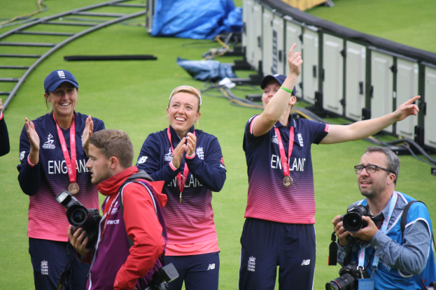 Group of people on a green field celebrating a victory, some wearing medals and caps, others holding cameras, with railings and bags in the background.