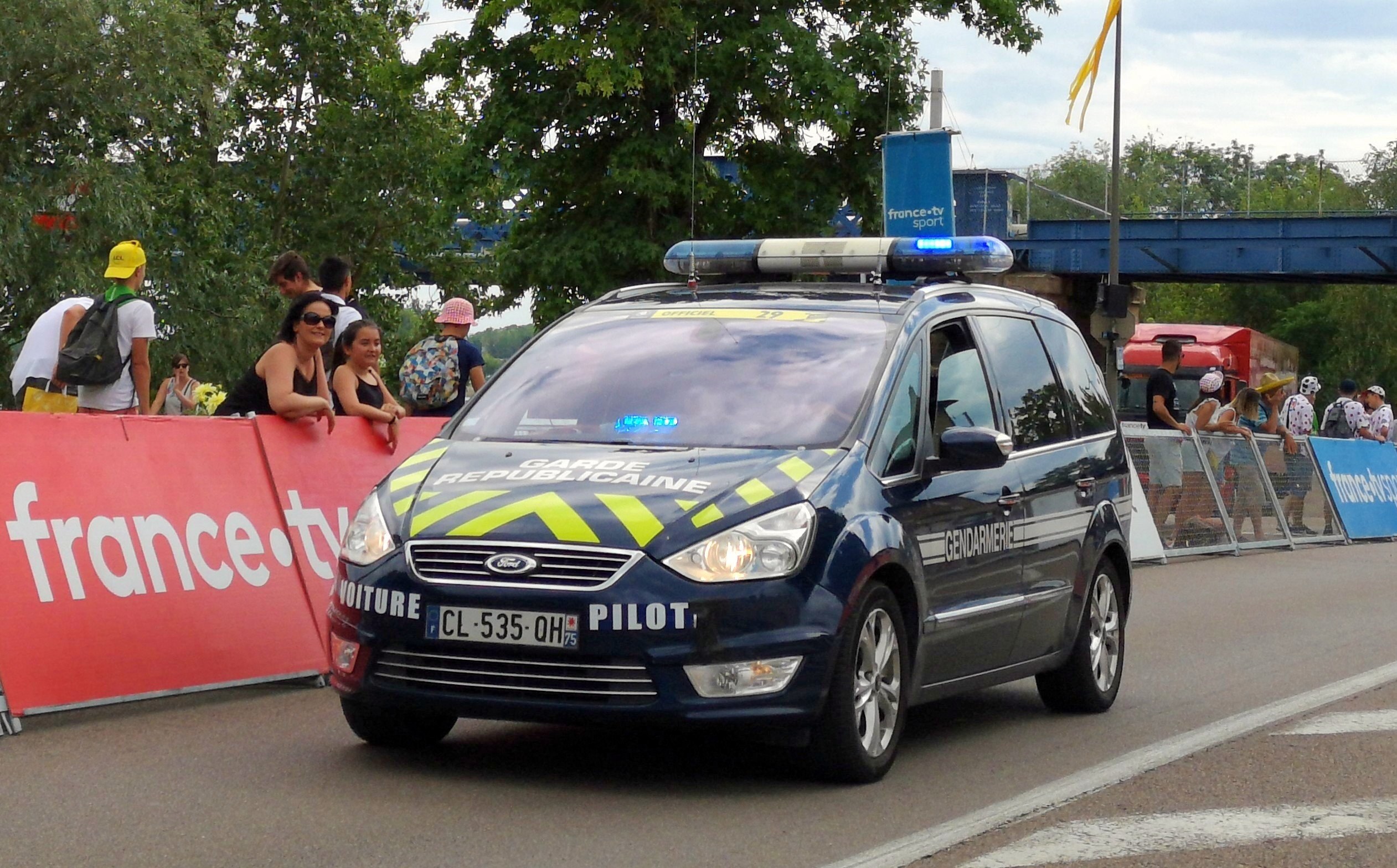 Polizeiauto fährt neben einer Menge auf einer Straße mit Bannern, Gelöndern, Bäumen, einer Brðcke, einer Flagge und einem bewölktem Himmel im Hintergrund.