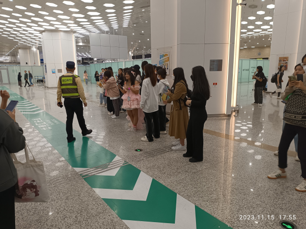 Eine Gruppe von Menschen, die in einer Reihe an einem Flughafen-Check-in-Schalter stehen, einige halten Taschen und Mobiltelefone, mit Säulen und Deckenleuchten im Hintergrund.