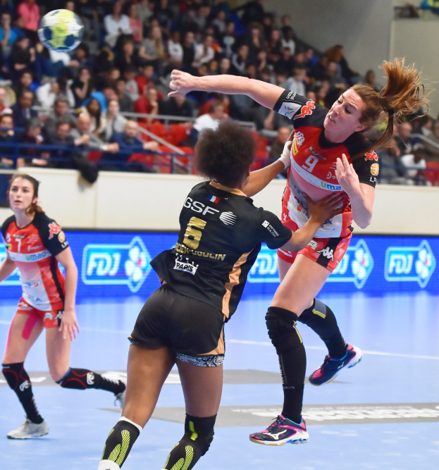 Eine Gruppe von Frauen spielt Handball auf einem Court mit einem Ball in der Luft, während Zuschauer von Stühlen, Geländern und einer Wand im Hintergrund zuschauen.