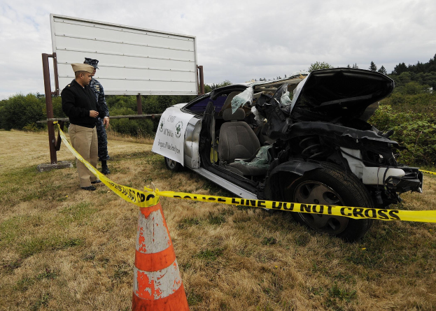 Ein Polizeibeamter in Uniform steht neben einem schwer beschädigten Auto am Straßenrand, mit eingedellter Motorhaube und zersplitterten Fenstern, in der Nähe eines Verkehrskegels und eines bandagierten Pfostens, mit Bäumen und einem bewölkten Himmel im Hintergrund.