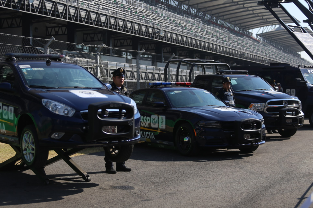 Polizeiautos vor einer Rennstrecke mit uniformierten Personen, im Hintergrund ein Stadion mit Geländern und Maschendrahtzaun.