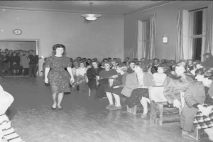 Eine Frau in einem schwarzen und weißen Kleid läuft über den Laufsteg während einer Modenschau für Frauenrechte, mit einer Menge, die auf Stühlen sitzt und einer Uhr und Lichtern an der Wand.