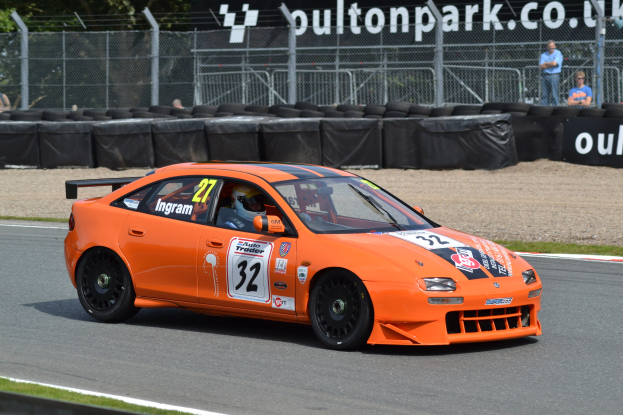 Oranges Auto auf einer Rennstrecke mit einem Helmträger am Steuer, umgeben von Reifen, Absperrungen, Zuschauern, Bäumen und einem Banner im Hintergrund.