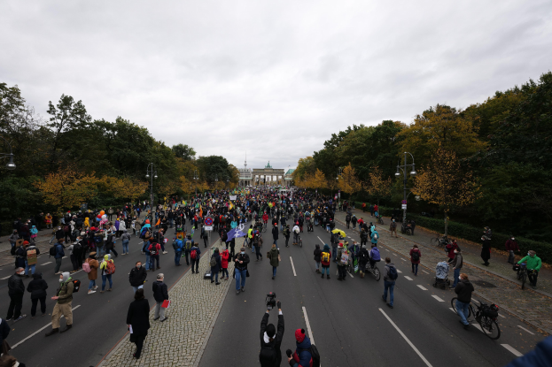 Eine große Gruppe von Menschen, die eine von Bäumen gesäumte Straße mit Laternenpfählen entlanggeht und Kameras hält, während sie an einer Protestdemo in Berlin teilnimmt, mit einem Gebäude und einem klaren Himmel im Hintergrund.