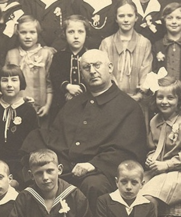 Ein Schwarz-Wei├č-Foto eines strengen Mannes, der in der Mitte sitzt und von einer Gruppe Kinder umgeben ist, die alle bewundernd zu ihm aufschauen.