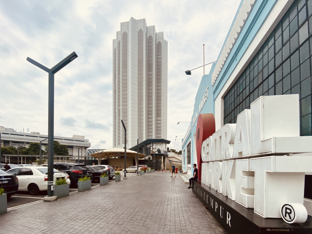 Ein belebter zentraler Markt in Kuala Lumpur, Malaysia, mit hohen Gebäuden, Straßenlaternen, Straßenlaternen, Kraftfahrzeugen, Fußgängern, Namensschildern, Bäumen und einem bewölkten Himmel.