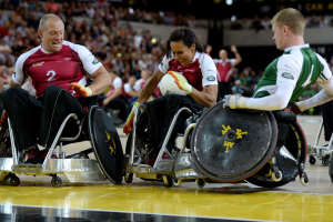 Eine Gruppe von Menschen in Rollstühlen spielt Handball mit einem Ball in der Mitte, mit unscharfen Zuschauern im Hintergrund.