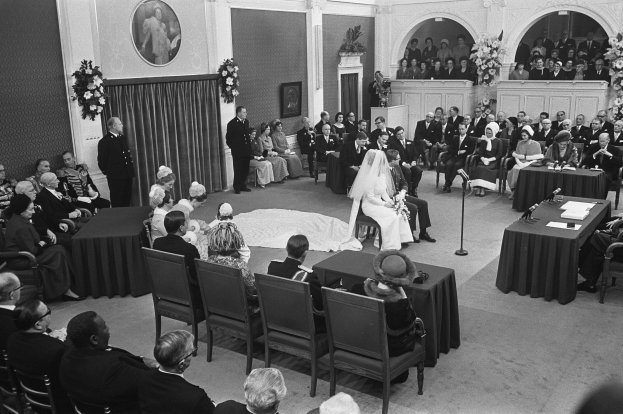 Ein Schwarz-Weiß-Foto einer Hochzeitszeremonie in einem großen Raum mit Gästen auf Stühlen und Stehplätzen, Tischen mit Papieren und Mikrofonen, einer dekorierten Wand mit Fotorahmen und Blumenbouquets und einem Podium mit Mikrofonständer.