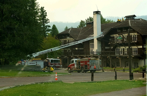 Feuerwehrfahrzeug vor einem großen Gebäude mit Fenstern, Geländern, Pfeilern und einem Schornstein geparkt, umgeben von Absperrböcken, Gras, Bäumen und Laternenmästen, mit einigen Menschen und Hügeln im Hintergrund unter einem klaren blauen Himmel.