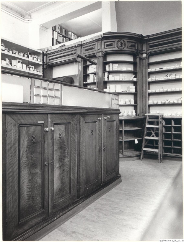 Schwarz-weiß-Foto eines vintage-Apothekeninneren mit einer Theke, Schränken, einer Leiter, Regalen voller Behälter und Text oben.