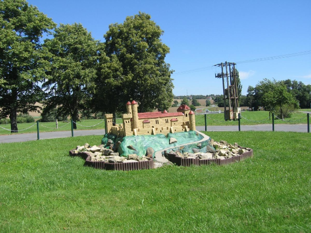 Minigolfplatz auf einer grünen Wiese, umgeben von einem Maschendrahtzaun, mit Bäumen, Strommasten und bewölktem Himmel im Hintergrund.