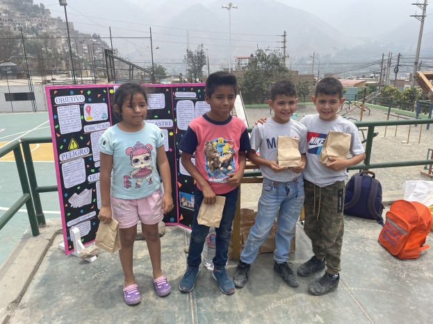 Gruppe von Kindern auf einem Basketballfeld, die Papierbeutel halten, mit Beuteln und Gegenständen rechts und einer Tafel mit Text links, vor einem Hintergrund aus Zaun, Pfählen, Bäumen, Gebäuden, Hügeln und Himmel.
