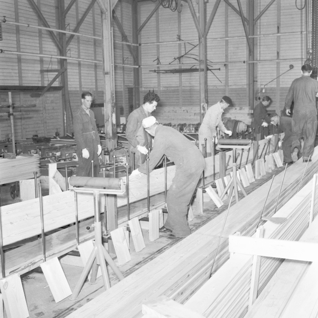 Schwarzes und weißes Bild von Männern, die in einer Fabrik mit Holzplanken auf dem Boden und Maschinen im Hintergrund arbeiten.