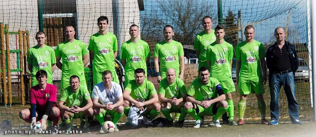 Eine Gruppe von Männern in grünen T-Shirts steht auf einem Fußballfeld, einige hocken, mit einem Ball vor ihnen in der Nähe eines Torpfostens, umgeben von Bäumen, Gebäuden und Fahrzeugen unter dem Himmel.
