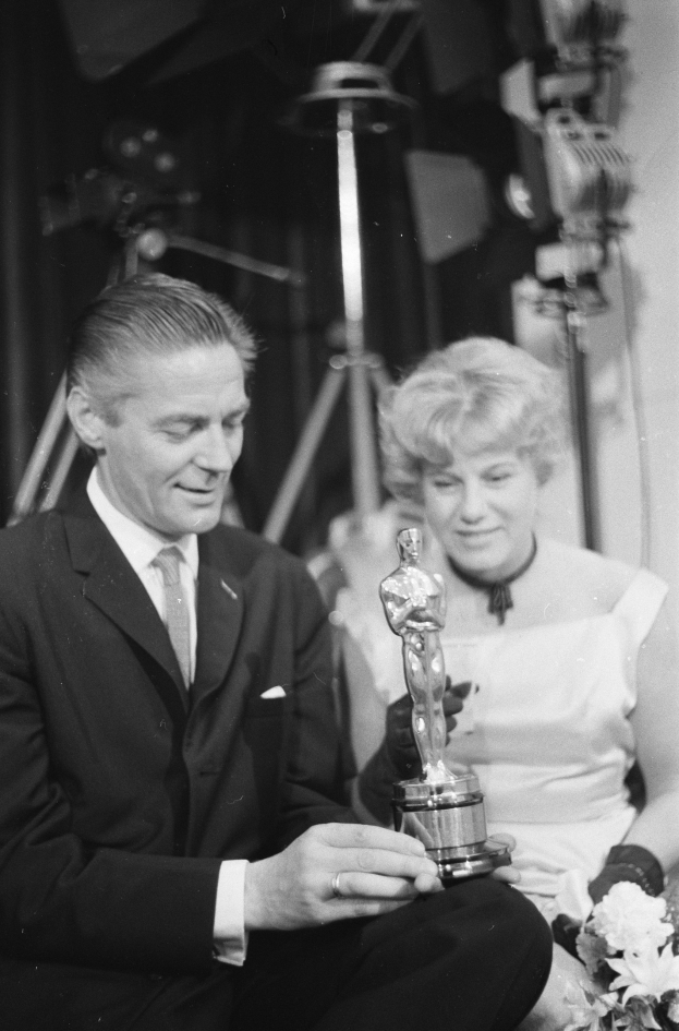 Ein Mann in einem schwarzen Blazer, weißem Hemd und Krawatte hölt eine Oscar-Statue, wa√hrend er neben einer Frau in Abendgarderobe sitzt, die einen Blumenstrauß hölt, mit Lichtern im Hintergrund.