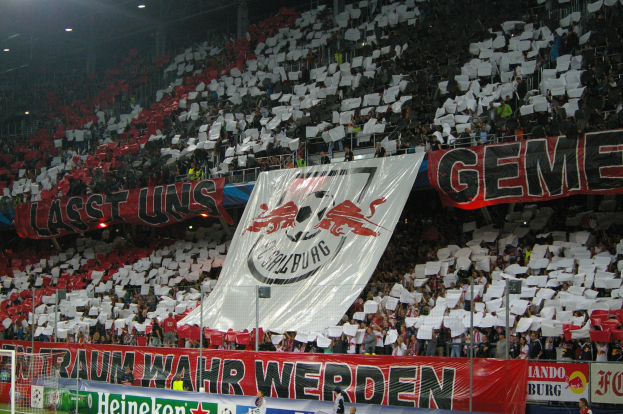 Ein überfülltes Fußballstadion mit Sitzplätzen und Stehplätzen, Banner mit Text und Logos, eine sichtbare Torstange und Überdachungsbeleuchtung.