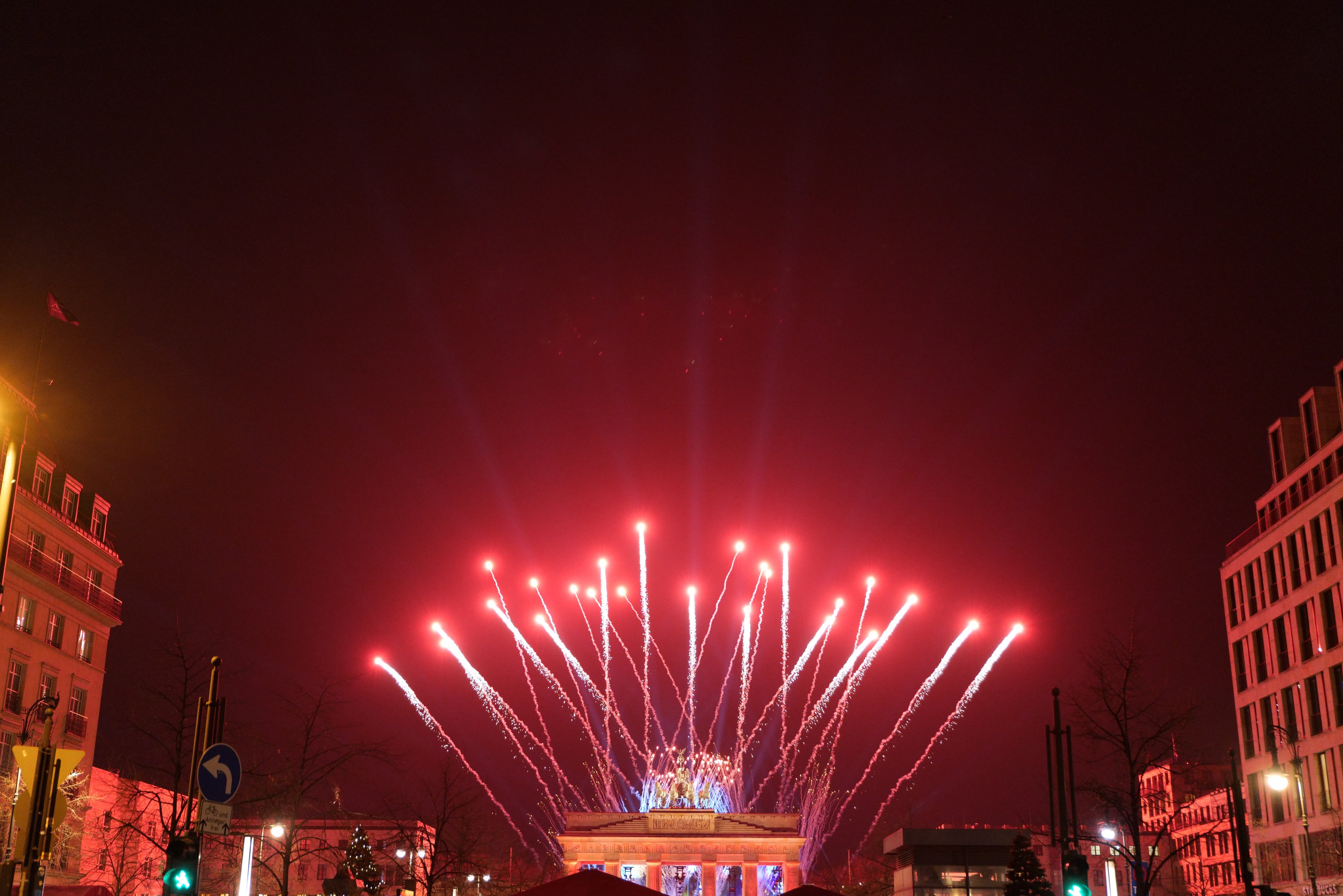 Nächtliche Ansicht des Berliner Brandenburger Tors mit Feuerwerk, das den Himmel über einer belebten Stadtstraße mit Gebäuden, Bäumen, Pfählen, Verkehrszeichen, Schildern und Flaggen erhellt.