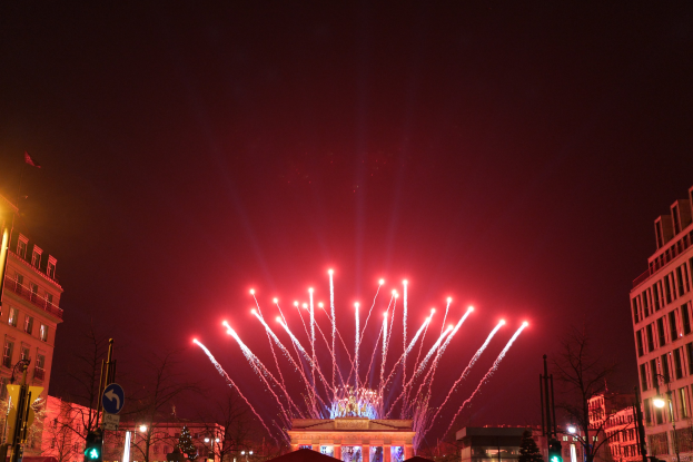 Nächtliche Ansicht des Berliner Brandenburger Tors mit Feuerwerk, das den Himmel über einer belebten Stadtstraße mit Gebäuden, Bäumen, Pfählen, Verkehrszeichen, Schildern und Flaggen erhellt.