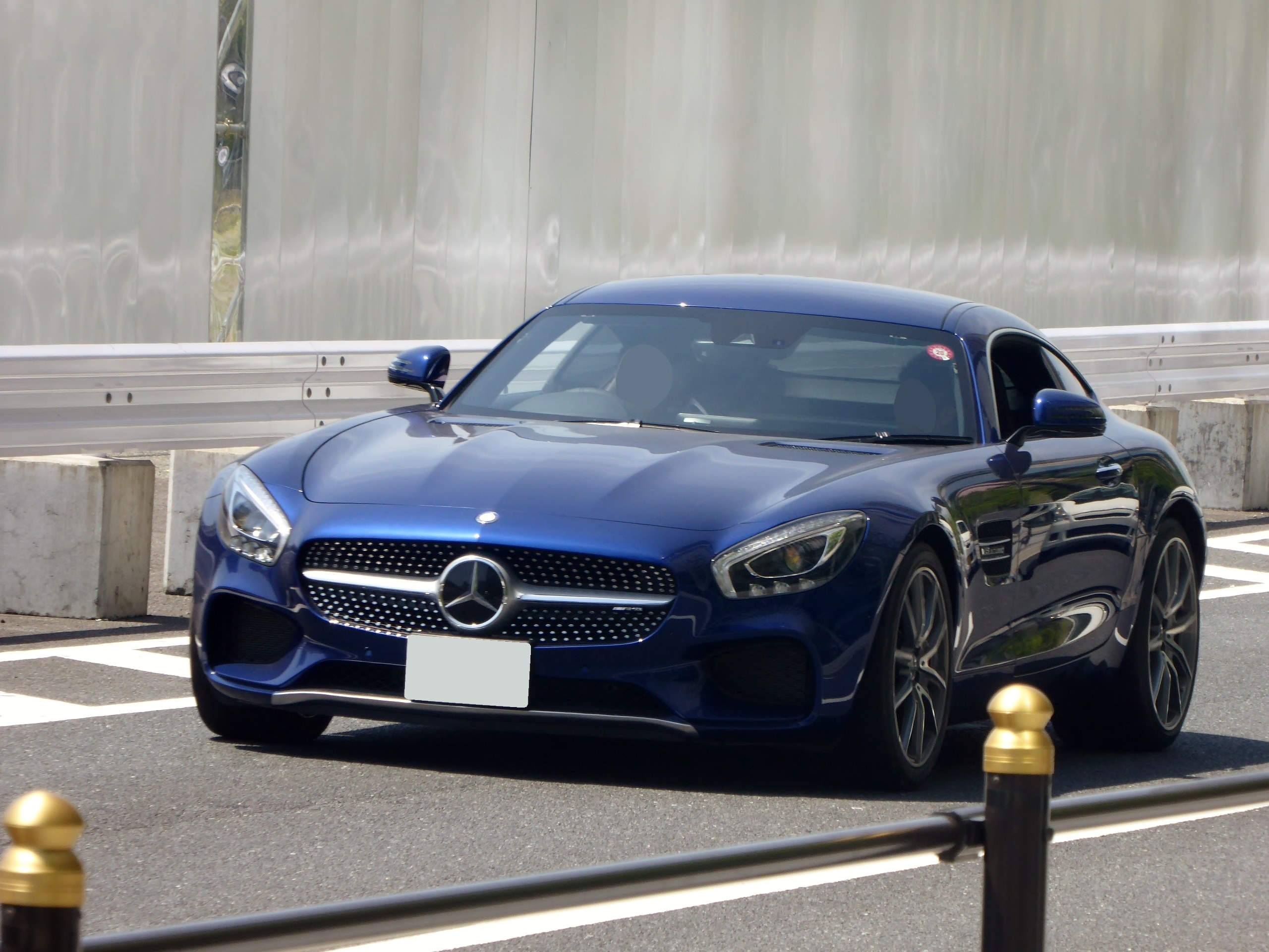 Ein blaues Mercedes-Benz AMG GT Coupé mit einer schlanken, aerodynamischen Gestaltung, eckigen Scheinwerfern, einem großen Kühlergrill, glänzenden Rädern und einem luxuriösen Lederinnenraum, das eine Straße mit einer Wand im Hintergrund entlangfährt.