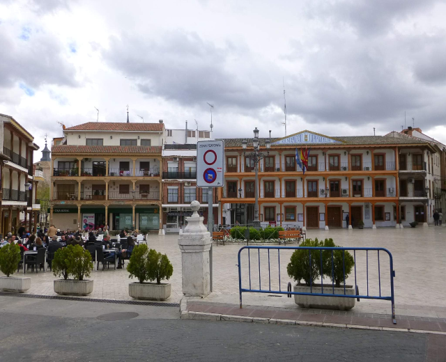 Ein belebter Stadtplatz mit sitzenden und stehenden Menschen, umgeben von Topfpflanzen, Metallabsperrungen, Schildern, Straßenlaternen mit Fahnen, Gebäuden und einem bewölkten Himmel.
