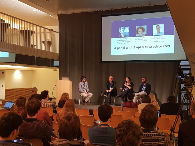 Eine Gruppe von Menschen sitzt in Stühlen gegenüber einem Projektionsschirm mit einem Laptop auf einem zentralen Tisch, eine Kamera auf einem Stativ rechts daneben und Geländern, Lampen und einer Wand im Hintergrund, die an einer Podiumsdiskussion über Open-Data-Aktivisten teilnehmen.