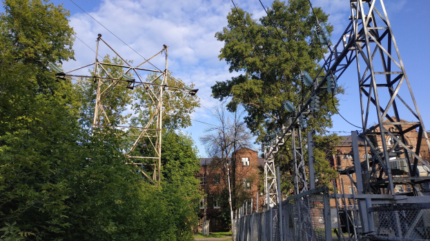 Gro├če Stromleitung durch einen bewaldeten Bereich mit Stützen, Dr├Ąhten und einem Zaun, mit im Hintergrund sichtbaren Geb├Ąuden unter einem bew├Âlktem Himmel.