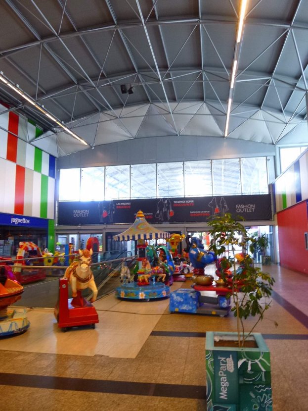 Amusement rides, a potted plant, scattered toys, informational boards, a wall, ceiling lights, and metal poles fill a large shopping mall room.