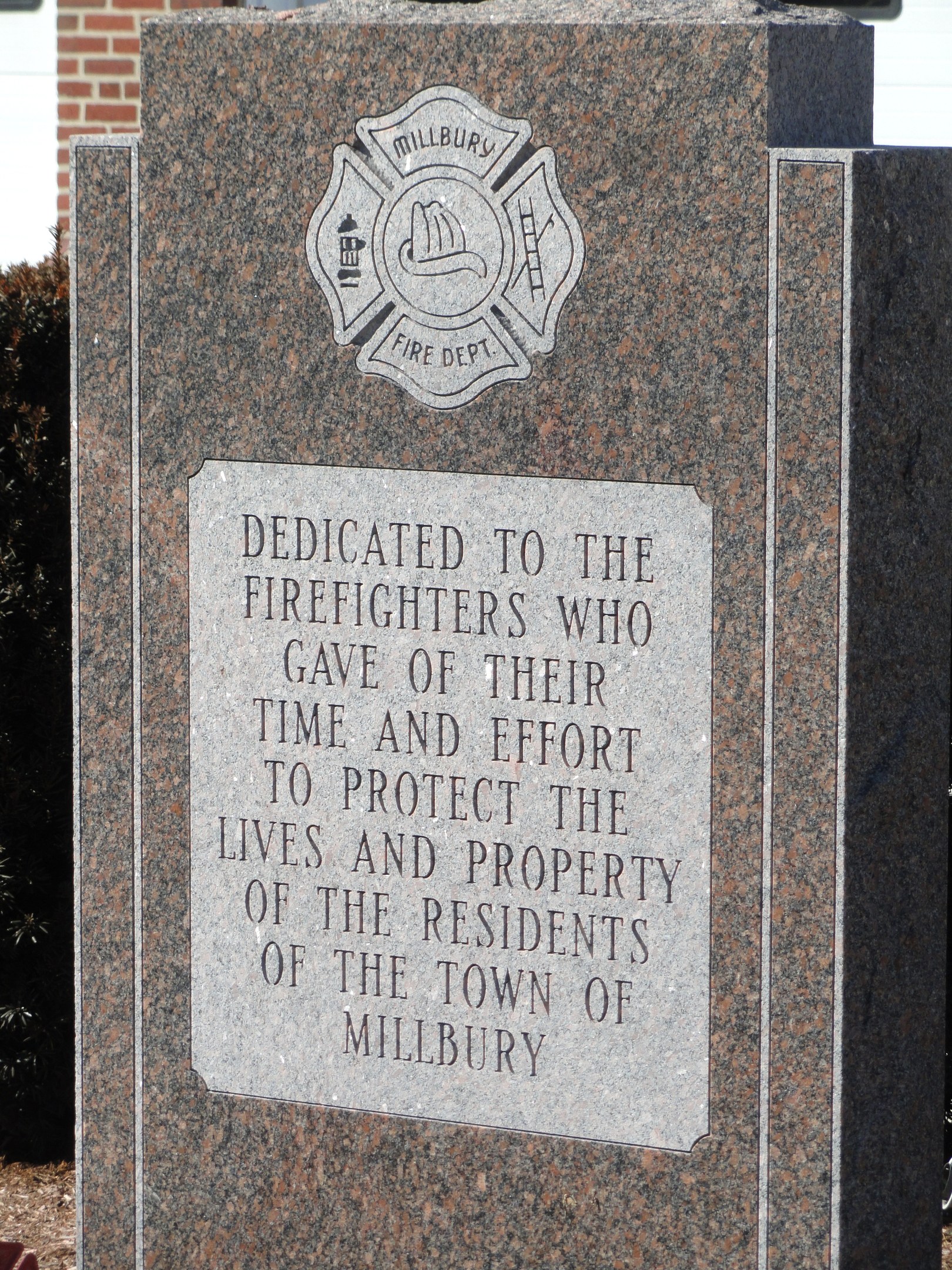 Denkmalstein mit einer Plakette, gewidmet den Feuerwehrleuten in Millbury, vor einem Backsteingebäude mit umgebender Vegetation.