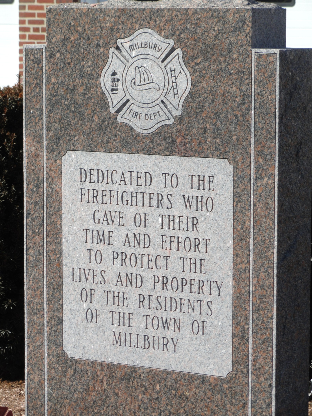 Denkmalstein mit einer Plakette, gewidmet den Feuerwehrleuten in Millbury, vor einem Backsteingebäude mit umgebender Vegetation.