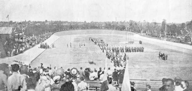 Schwarzes und weißes Foto einer Menge, die ein Pferderennen in einem Stadion beobachtet, mit Zuschauern auf Bänken und Boden, einige halten Fahnen und andere reiten Pferde, vor einem Hintergrund aus Bäumen und einem klaren Himmel.