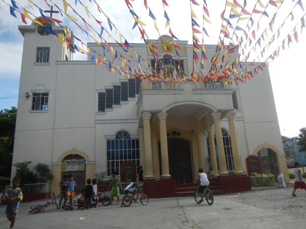 Eine Gruppe von Menschen, die Fahrräder vor einer weißen Kirche fahren, mit architektonischen Details wie Fenstern, Säulen, Bögen, Türen und einem Kreuzsymbol, mit Pflanzen, einem Zaun, einer Fahne, Bäumen und einem bewölkten Himmel im Hintergrund und bunten Fahnen, die an einem Seil oben befestigt sind.