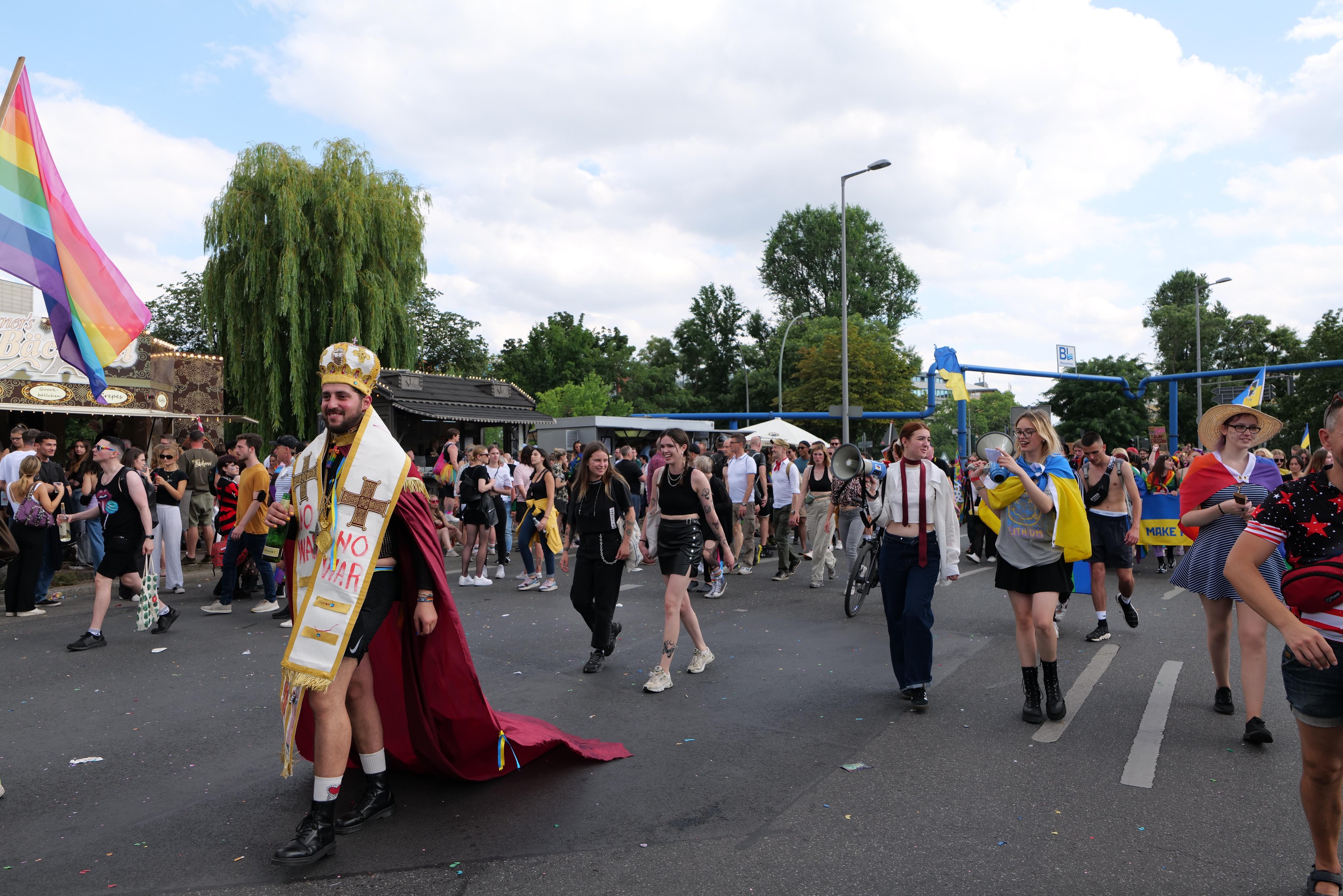 Eine Gruppe von Menschen marschiert bei der Pride Parade 2018 mit einer Regenbogenfahne und Musikinstrumenten, umgeben von Laternenmasten, Bäumen, Hütten und einem bewölkten Himmel.