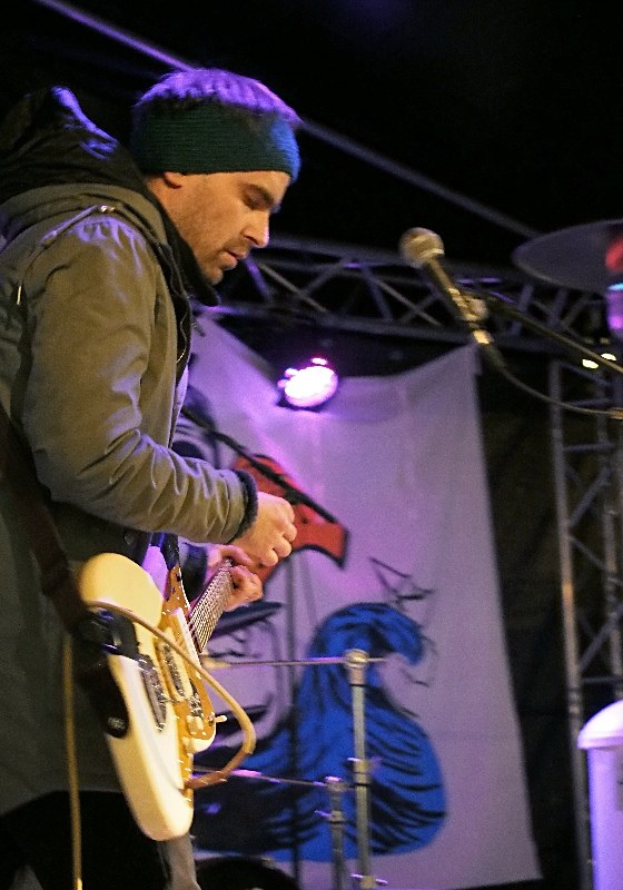 Ein Mann in einer Jacke und einer Mütze spielt eine E-Gitarre auf der Bühne, mit einem Mikrofon davor, einem Schlagzeug zu seiner Rechten und beleuchteten Bannern im Hintergrund.
