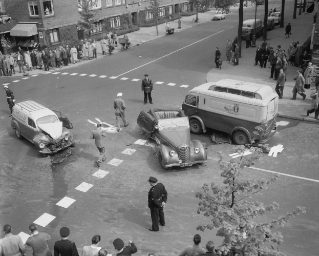 Schwarz-weiß-Szene eines Autounfalls an einer Straßenkreuzung mit mehreren Fahrzeugen, darunter ein Lieferwagen, und einer Gruppe von Menschen, die danebenstehen, mit Gebäuden, Bäumen, Pfosten und Texttafeln im Hintergrund.