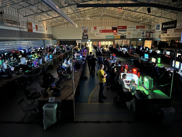 Ein großer Raum mit vielen Menschen, die Videospiele spielen, sitzen und stehen, umgeben von Monitoren, Tastaturen, Mäusen und Kabeln auf Tischen, mit markierten Platten im Hintergrund und sichtbarer Decke.