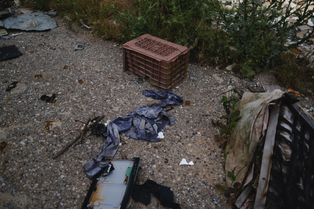 Müllhaufen einschließlich einer Kunststoffbox und Kleidung auf der Seite einer Straße in der Nähe eines Zauns, mit Pflanzen und Gras im Hintergrund.
